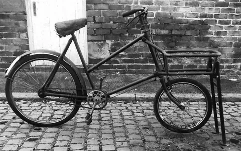 Black and white photo of a carrier bike with a big front rack, against a brick wall