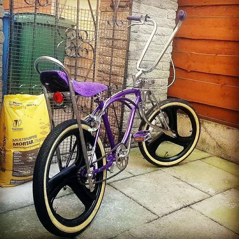 Purple lowrider with tall chrome bars, a sissy bar, a purple saddle and three-spoke wheels