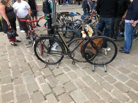 A black roadster and a bike with a large wicker basket, on display stands on cobbles